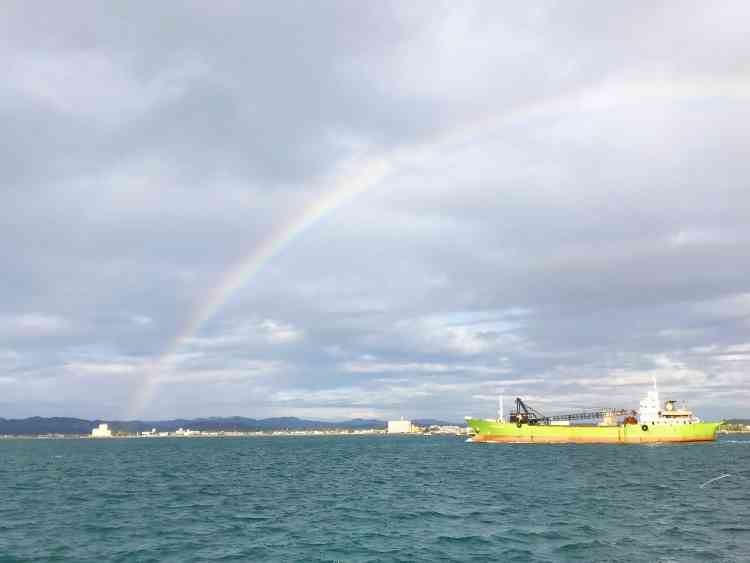 雨上がりと共に、海の上に虹が掛かっている様子。右側には船に浮かぶ貨物船が見える。